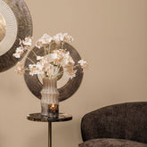 Wandpaneel Jacci Goud Rond Small van PTMD, paneel deels achter een vaas met witte bloesem en een donkere fauteuil