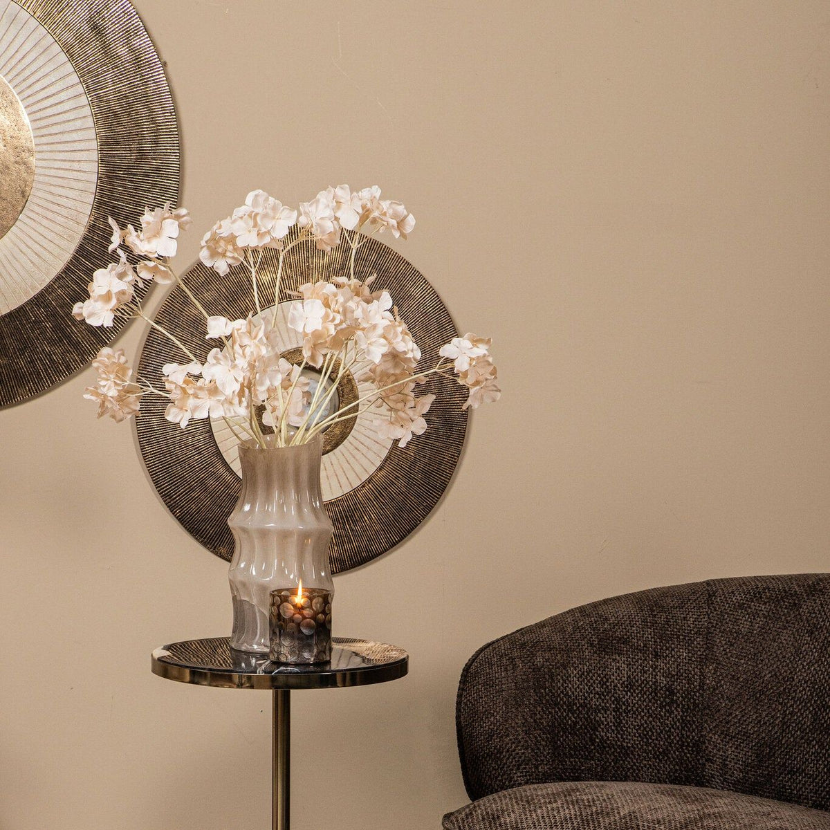 Wandpaneel Jacci Goud Rond Small van PTMD, paneel deels achter een vaas met witte bloesem en een donkere fauteuil