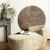 Wandpaneel Wiktor Rond Bruin Ø120cm van PTMD in een lichte woonkamer met beige fauteuil en hocker